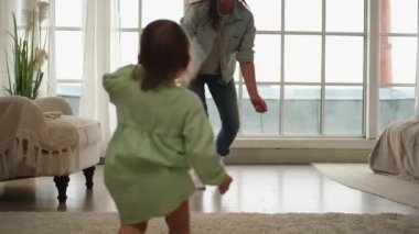 Bebek gelişimi. Evde ilk adımlarını atan tatlı küçük bir kız. Oturma odasında yürüyen sevimli bebeğe bakarak gülümseyen mutlu moter. Evde mutlu bir aile. Anne bebeğe sevgiyle sarılıyor..