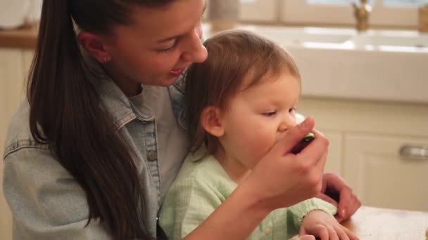 快乐的家庭在家里妈妈在厨房里用勺子喂女儿 小孩子的脸很乱 很滑稽 在家里吃着健康的食物 给小女儿喂食的年轻妈妈 — 图库视频影像