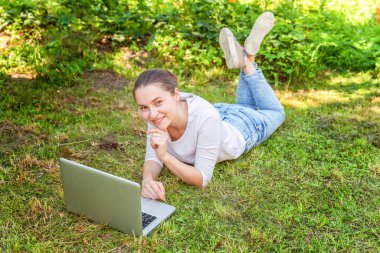 Serbest iş konsepti. Genç kadın dizüstü bilgisayar üzerinde çalışan şehir parkında yeşil çim çim üzerinde yatan. Yaşam tarzı otantik samimi öğrenci kız açık havada eğitim. Mobil Ofis
