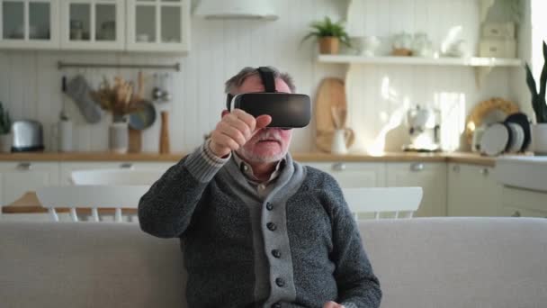 自宅でVrメガネのヘッドセットを仮想現実を使って身に着けている興奮した熟年男性 バーチャルリアリティヘルメットのVr体験中に祖父が空気に触れる シミュレーションハイテクビデオゲーム — ストック動画
