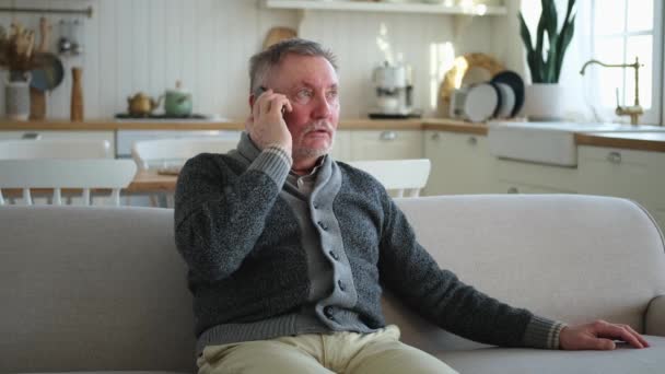 stock video Happy middle aged senior man talking on smartphone with family friends. Older mature grandfather with cell phone talking with grown up children, resting at home. Older generation modern tech usage