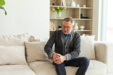 Diz ağrısı, eklem iltihabı, vücut hastalıkları, sağlık hizmetleri. Evdeki kanepede oturan mutsuz, orta yaşlı, diz ağrısı çeken bir adam. Yaşlı büyükbaba bacağına dokunuyor ve dizinde ağrı hissediyor.