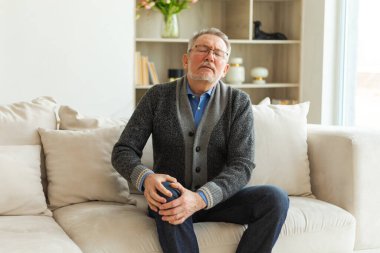 Diz ağrısı, eklem iltihabı, vücut hastalıkları, sağlık hizmetleri. Evdeki kanepede oturan mutsuz, orta yaşlı, diz ağrısı çeken bir adam. Yaşlı büyükbaba bacağına dokunuyor ve dizinde ağrı hissediyor.