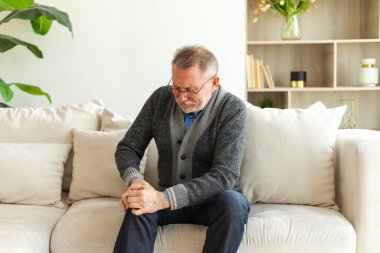 Diz ağrısı, eklem iltihabı, vücut hastalıkları, sağlık hizmetleri. Evdeki kanepede oturan mutsuz, orta yaşlı, diz ağrısı çeken bir adam. Yaşlı büyükbaba bacağına dokunuyor ve dizinde ağrı hissediyor.