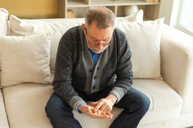 Evdeki kanepede oturan orta yaşlı bir adam elinde tıbbi haplar tutuyor. Yaşlı büyükbaba ilaç tedavisi hapı vitamini alıyor. Yaş reçeteli ilaç sağlık tedavisi kavramı