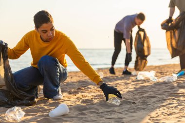 Dünya günü. Gönüllü eylemciler sahil bölgesindeki çöpleri topluyor. Kadınlar ve erkekler okyanus kıyısındaki çöp torbasına plastik çöp koyar. Çevre koruma kıyı bölgesi temizliği