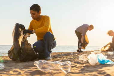 Dünya günü. Gönüllü eylemciler sahil bölgesindeki çöpleri topluyor. Kadınlar ve erkekler okyanus kıyısındaki çöp torbasına plastik çöp koyar. Çevre koruma kıyı bölgesi temizliği