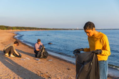 Dünya günü. Gönüllü eylemciler sahil bölgesindeki çöpleri topluyor. Kadınlar ve erkekler okyanus kıyısındaki çöp torbasına plastik çöp koyar. Çevre koruma kıyı bölgesi temizliği