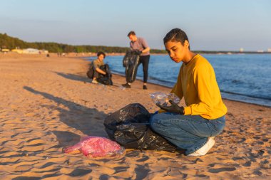 Dünya günü. Gönüllü eylemciler sahil bölgesindeki çöpleri topluyor. Kadınlar ve erkekler okyanus kıyısındaki çöp torbasına plastik çöp koyar. Çevre koruma kıyı bölgesi temizliği