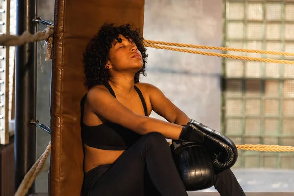 Woman fighter girl power. African american woman fighter with boxing gloves sitting on boxing ring waiting and resting after fight. Strong powerful girl in gym. Strength fit body workout training