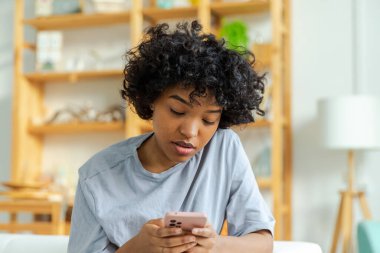 African girl holding smartphone touch screen typing scroll page at home. Woman with cell phone surfing internet using social media apps playing game. Shopping online Internet news cellphone addiction