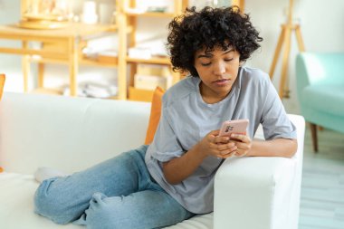 African girl holding smartphone touch screen typing scroll page at home. Woman with cell phone surfing internet using social media apps playing game. Shopping online Internet news cellphone addiction