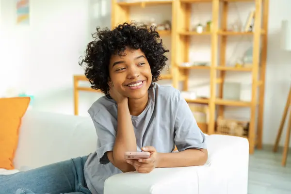 stock image African girl holding smartphone touch screen typing scroll page at home. Woman with cell phone surfing internet using social media apps playing game. Shopping online Internet news cellphone addiction