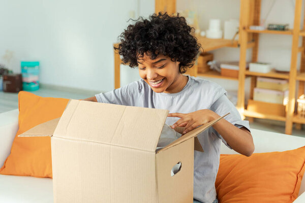African girl unpacking delivery looking in box. Happy woman opening carton box. Female getting parcel looking at delivered goods items. Satisfied client positive feedback quick express deliver