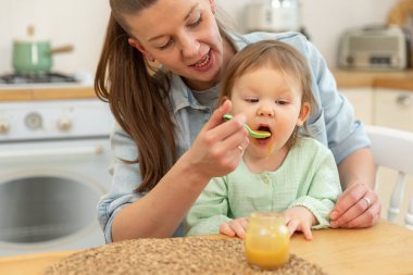 Evde mutlu bir aile. Anne mutfakta bebeğini kaşıkla besliyor. Dağınık suratlı küçük çocuk evde sağlıklı yiyecekler yiyor. Genç kadın anne, küçük kızına yemek veriyor.