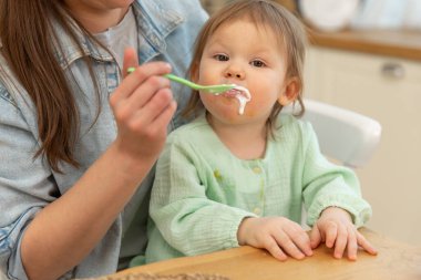 Evde mutlu bir aile. Anne mutfakta bebeğini kaşıkla besliyor. Dağınık suratlı küçük çocuk evde sağlıklı yiyecekler yiyor. Genç kadın anne, küçük kızına yemek veriyor.