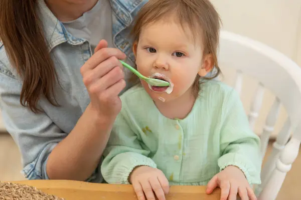 Happy family at home. Mother feeding her baby girl from spoon in kitchen. Little toddler child with messy funny face eats healthy food at home. Young woman mom giving food to kid daughter