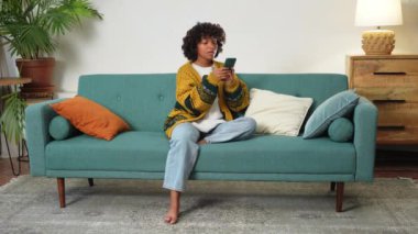 African girl holding smartphone touch screen typing scroll page at home. Woman with cell phone surfing internet using social media apps playing game. Shopping online Internet news cellphone addiction