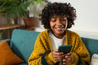 African girl holding smartphone touch screen typing scroll page at home. Woman with cell phone surfing internet using social media apps playing game. Shopping online Internet news cellphone addiction