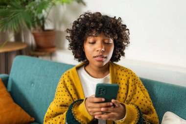 African girl holding smartphone touch screen typing scroll page at home. Woman with cell phone surfing internet using social media apps playing game. Shopping online Internet news cellphone addiction