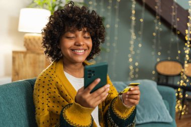 African american woman shopping online holding smartphone paying with gold credit card. Girl sitting at home buying on Internet enter credit card details. Online shopping ecommerce delivery service
