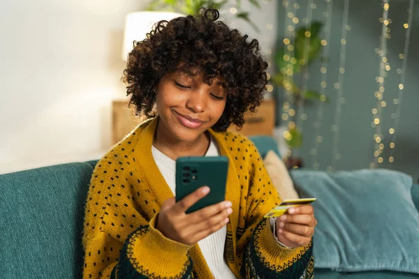 African american woman shopping online holding smartphone paying with gold credit card. Girl sitting at home buying on Internet enter credit card details. Online shopping ecommerce delivery service