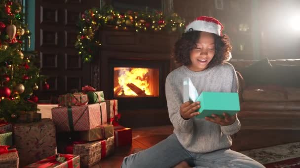 メリークリスマス アフリカ系アメリカ人女性がクリスマスツリーの近くにギフトボックスを開けます クリスマスツリーと暖炉付きのリビングルームの少女はサプライズフェイスでギフトボックスを開きます 自宅でクリスマスイブ — ストック動画