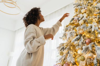 Mutlu noeller. Noel ağacı süsleyen Afrikalı Amerikalı kadın. Klasik Noel ağacının yanında beyaz gümüş süslemeleri olan mutlu bir kız. Noel arifesinde evde kutlama zamanı.