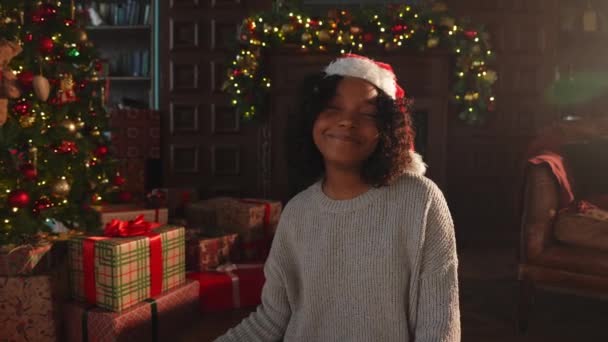 メリークリスマス アフリカ系アメリカ人女性は クラシックな暗いインテリアでクリスマスツリーの近くで微笑んでいます 暖炉のクリスマスツリーギフトボックス付きのリビングルームでサンタハットで幸せな少女 自宅でクリスマスイブ — ストック動画