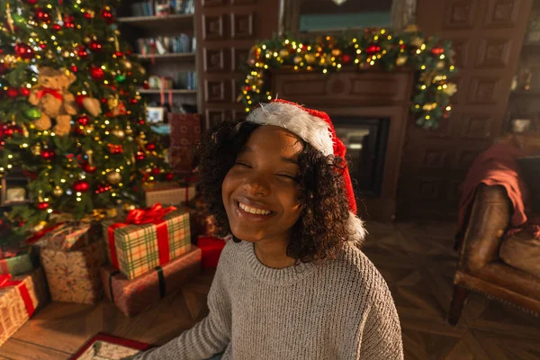 Merry Christmas. African American woman smiling near Christmas tree in classical dark interior. Happy girl in Santa hat in living room with fireplace Christmas tree gift boxes. Christmas eve at home