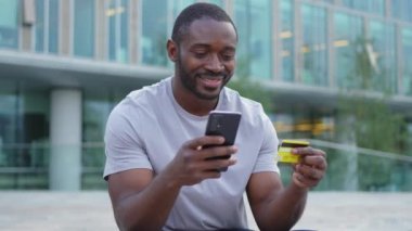 Çevrimiçi alışveriş yapan Afro-Amerikalı adam elinde altın kredi kartıyla alışveriş yapıyor. Şehirdeki sokaktaki adam internet üzerinden alıyor. Kredi kartı detaylarını gir. Online alışveriş ekommerce dağıtım servisi.