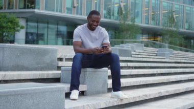 Şehirde akıllı telefonlu mutlu Afrikalı Amerikalı adam mutluluktan havalara uçuyor. Cep telefonuna bakan adam harika haberler okuyor ve online ihaleyi kazanırken çok şaşırıyor. Kazanan jest