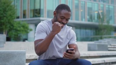 Şehirde akıllı telefonlu mutlu Afrikalı Amerikalı adam mutluluktan havalara uçuyor. Cep telefonuna bakan adam harika haberler okuyor ve online ihaleyi kazanırken çok şaşırıyor. Kazanan jest