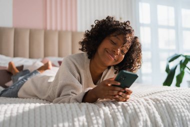 African girl holding smartphone touch screen typing scroll page at home. Woman with cell phone surfing internet using social media apps playing game. Shopping online Internet news cellphone addiction