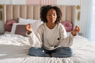 Yoga farkındalığı meditasyonu. Genç sağlıklı Afrikalı kız evde yoga yapıyor. Nilüfer çiçekli bir kadın yatakta oturmuş, meditasyon yapıyor, gülümsüyor, evde dinleniyor. Nefes egzersizi yapan bir kız. Evde yoga.