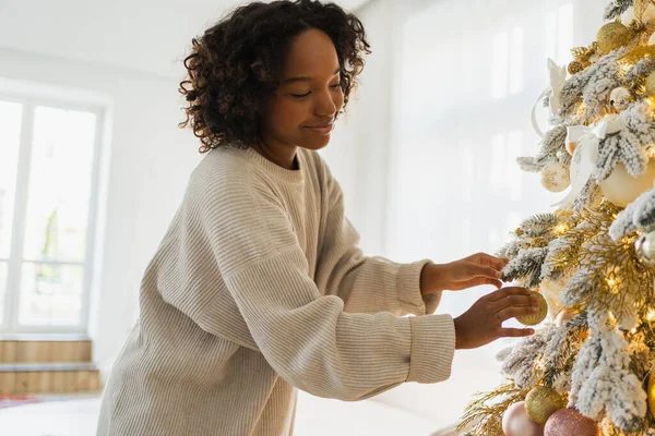 Merry Christmas. African American woman decorating Christmas tree. Happy girl near classical Christmas tree with white golden silver decorations ornament. Christmas eve at home time for celebration