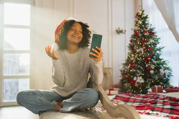 Merry Christmas. African woman in Santa hat having video chat in smartphone. Blogger in festive mood recording video in social network. Girl having video call with friends family celebrating Christmas