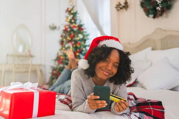 African American woman shopping online holding smartphone paying with gold credit card for Christmas gifts. Girl buying on Internet enter credit card details enjoying Christmas winter holidays at home