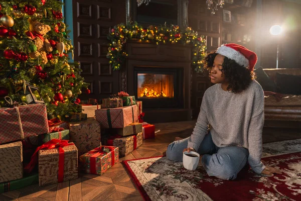 Merry Christmas. African American woman in Santa hat with cup of hot drink coffee tea near Christmas tree in dark interior. Girl in living room with Christmas tree and fireplace. Christmas eve at home
