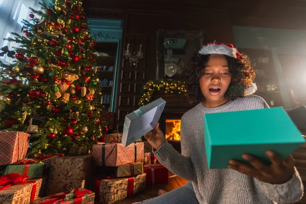 Merry Christmas. African American woman unpacking open up gift box near Christmas tree. Girl in living room with Christmas tree and fireplace opening gift box with surprise face. Christmas eve at home