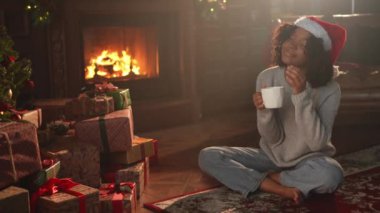 Mutlu noeller. Noel Baba şapkalı Afro-Amerikalı kadın, koyu renkli bir Noel ağacının yanında bir fincan sıcak kahve çayıyla. Oturma odasında Noel ağacı ve şöminesi olan bir kız. Evde Noel arifesi