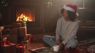 Mutlu noeller. Noel Baba şapkalı Afro-Amerikalı kadın, koyu renkli bir Noel ağacının yanında bir fincan sıcak kahve çayıyla. Oturma odasında Noel ağacı ve şöminesi olan bir kız. Evde Noel arifesi