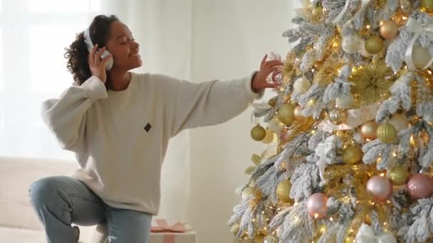 メリークリスマス アフリカ系アメリカ人女性は クリスマスツリーを飾る音楽を聴くヘッドフォンを着用しています 伝統的なクリスマスツリーに近いハッピーガール お祝いのための自宅時間でのクリスマスイブ — ストック動画