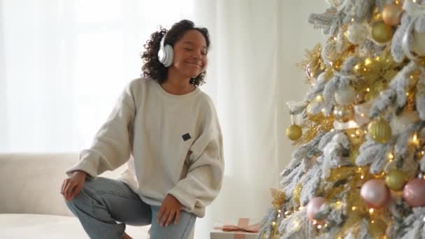 メリークリスマス アフリカ系アメリカ人女性は クリスマスツリーを飾る音楽を聴くヘッドフォンを着用しています 伝統的なクリスマスツリーに近いハッピーガール お祝いのための自宅時間でのクリスマスイブ — ストック動画
