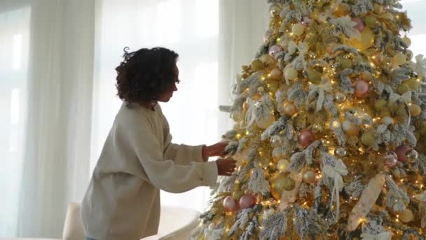 メリークリスマス クリスマスツリーを飾るアフリカ系アメリカ人女性 ホワイトゴールドシルバーの装飾の古典的なクリスマスツリーに近いハッピーガール お祝いのための自宅時間でのクリスマスイブ — ストック動画