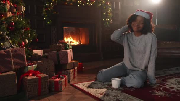 メリークリスマス サンタハットのアフリカ系アメリカ人女性は 暗いインテリアでクリスマスツリーの近くでホットドリンクコーヒーティーを飲みます クリスマスツリーと暖炉のあるリビングルームの女の子 自宅でクリスマスイブ — ストック動画