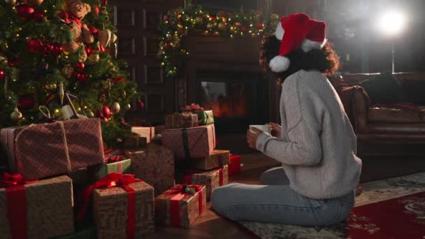 メリークリスマス アフリカ系アメリカ人女性は 古典的な暗いインテリアのクリスマスツリーの近くで暖炉を見ています クリスマスツリーギフトボックス付きのリビングルームのサンタハットの少女 自宅でクリスマスイブ — ストック動画