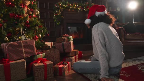 メリークリスマス アフリカ系アメリカ人女性は 古典的な暗いインテリアのクリスマスツリーの近くで暖炉を見ています クリスマスツリーギフトボックス付きのリビングルームのサンタハットの少女 自宅でクリスマスイブ — ストック動画