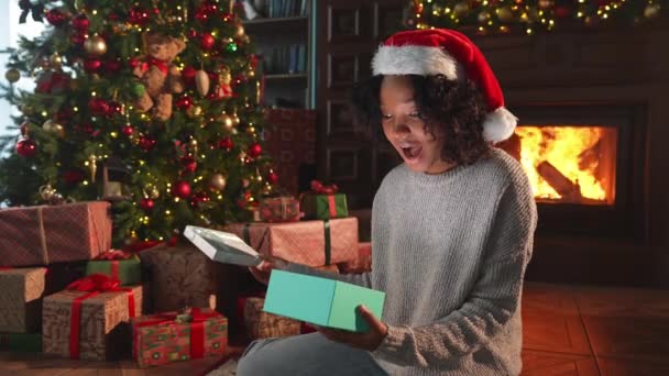 メリークリスマス アフリカ系アメリカ人女性がクリスマスツリーの近くにギフトボックスを開けます クリスマスツリーと暖炉付きのリビングルームの少女はサプライズフェイスでギフトボックスを開きます 自宅でクリスマスイブ — ストック動画