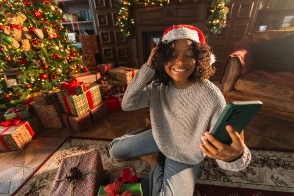 Merry Christmas. African woman in Santa hat having video chat in smartphone. Blogger in festive mood recording video in social network. Girl having video call with friends family celebrating Christmas